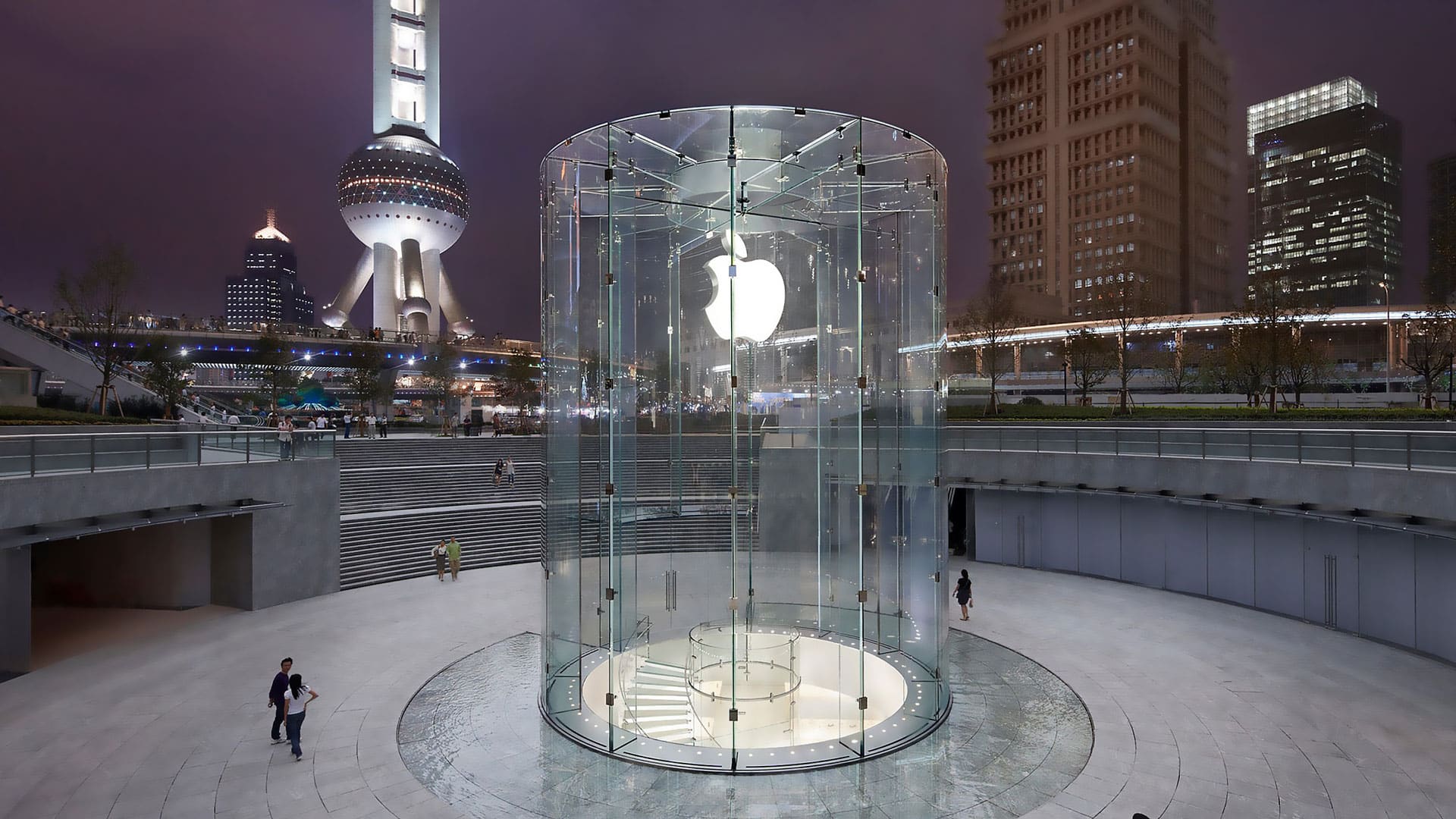 A cylindrical glass entrance featuring the Apple logo leads to an underground Apple Store in a cityscape with tall, illuminated buildings and the iconic Oriental Pearl Tower in the background at night. Amid a sales surge due to iPhone discounts in China, a few people are walking near the entrance.