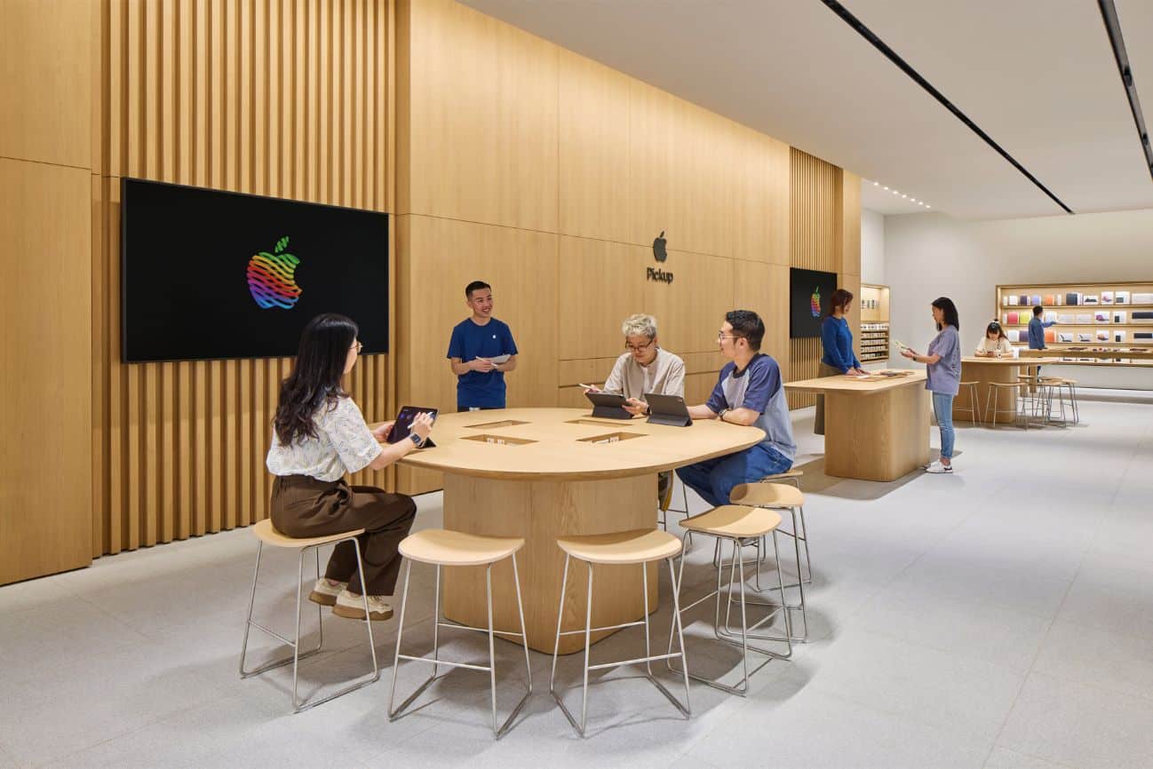 Nội thất cửa hàng Apple hiện đại, tràn ngập ánh sáng với các tấm và bàn gỗ ở Trung Quốc. Bốn khách hàng ngồi ở một chiếc bàn tròn, đang sử dụng các thiết bị của Apple trong khi một nhân viên đứng gần đó. Các kệ trưng bày sản phẩm xếp dọc theo bức tường, phản ánh doanh số bán hàng tăng vọt nhờ đợt giảm giá iPhone gần đây.