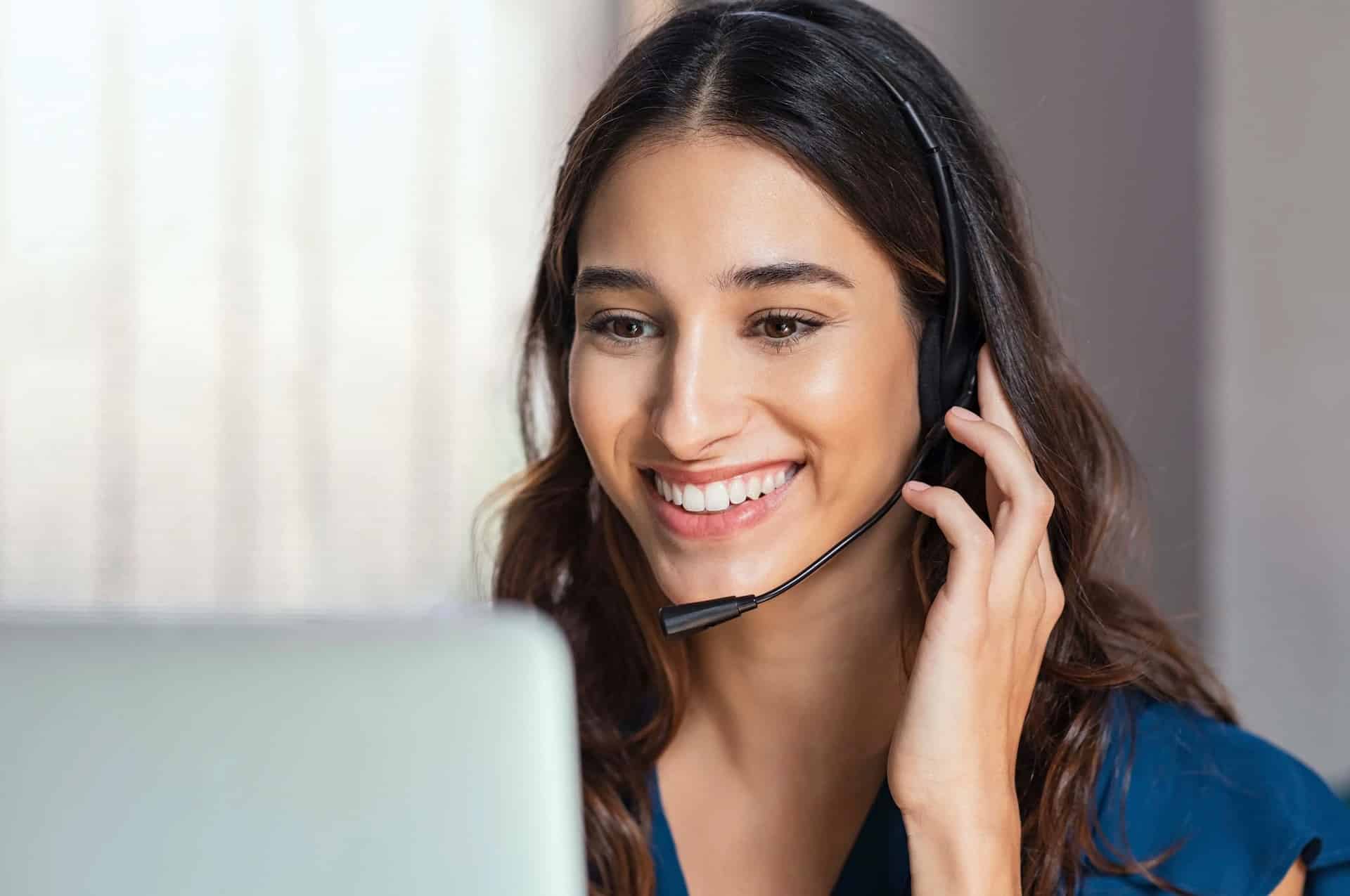 A woman wearing a headset and smiling while looking at a laptop. She has long brown hair and is seated indoors, possibly engaged in a video call or online conversation. The background is softly blurred, focusing attention on her face, reflecting her role in Customized Call Management Services.