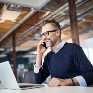 A man with glasses and a beard is sitting at a desk in an office, talking on a mobile phone. He is smiling and looking at a laptop in front of him, likely discussing customized call management services. The office has a modern design with exposed ducts and large windows.