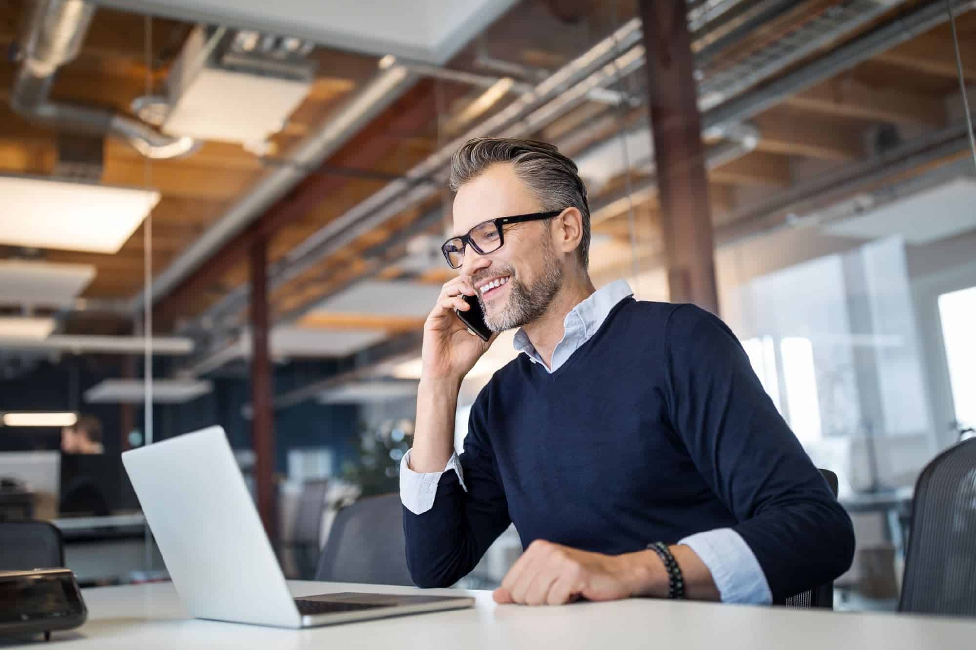 A man with glasses and a beard is sitting at a desk in an office, talking on a mobile phone. He is smiling and looking at a laptop in front of him, likely discussing customized call management services. The office has a modern design with exposed ducts and large windows.