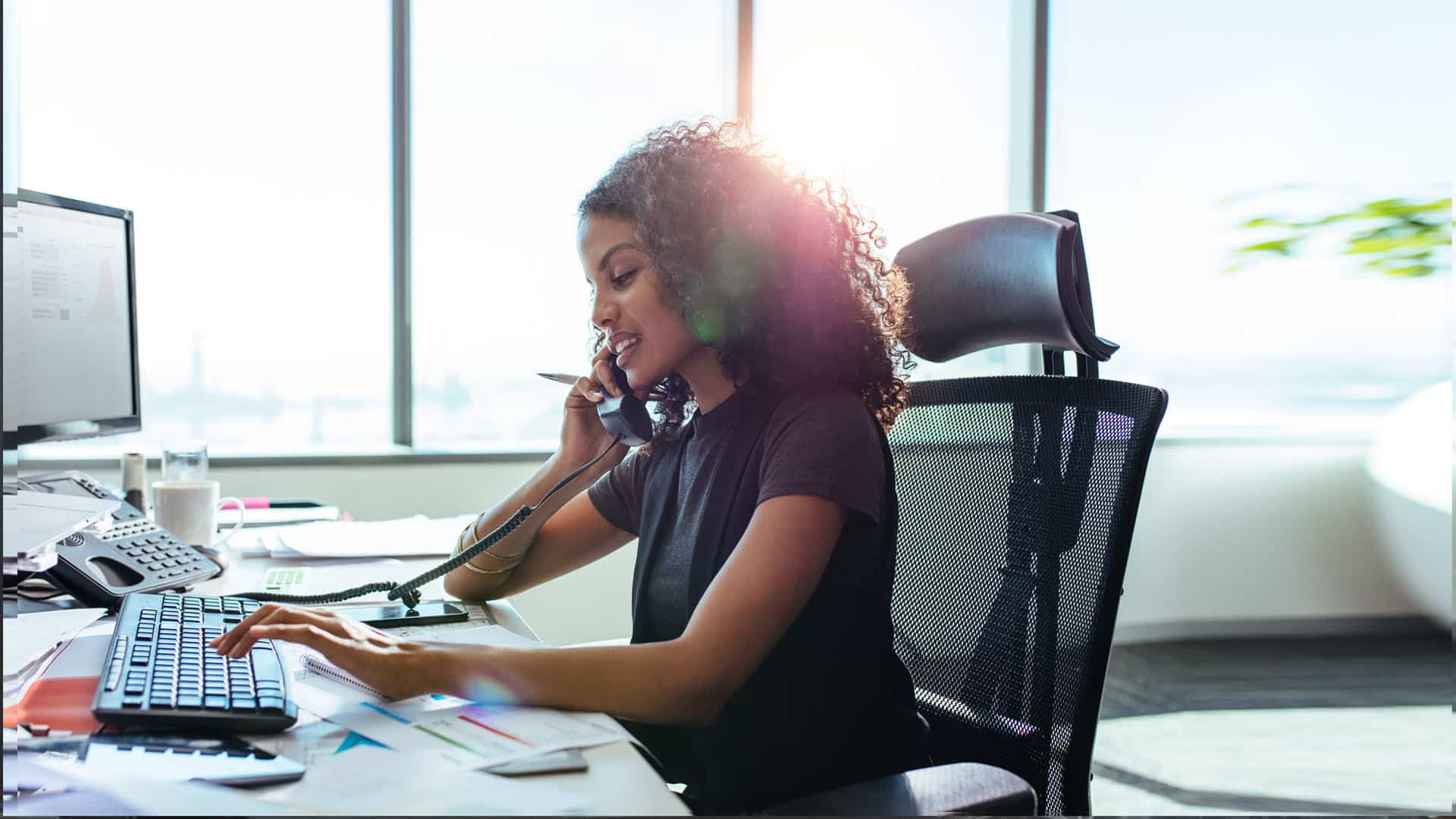 A woman with curly hair sits at an office desk, talking on the phone about Customized Call Management Services. She types on a computer keyboard and looks focused. Documents and office supplies are scattered on the desk, and there are large windows in the background letting in bright sunlight.