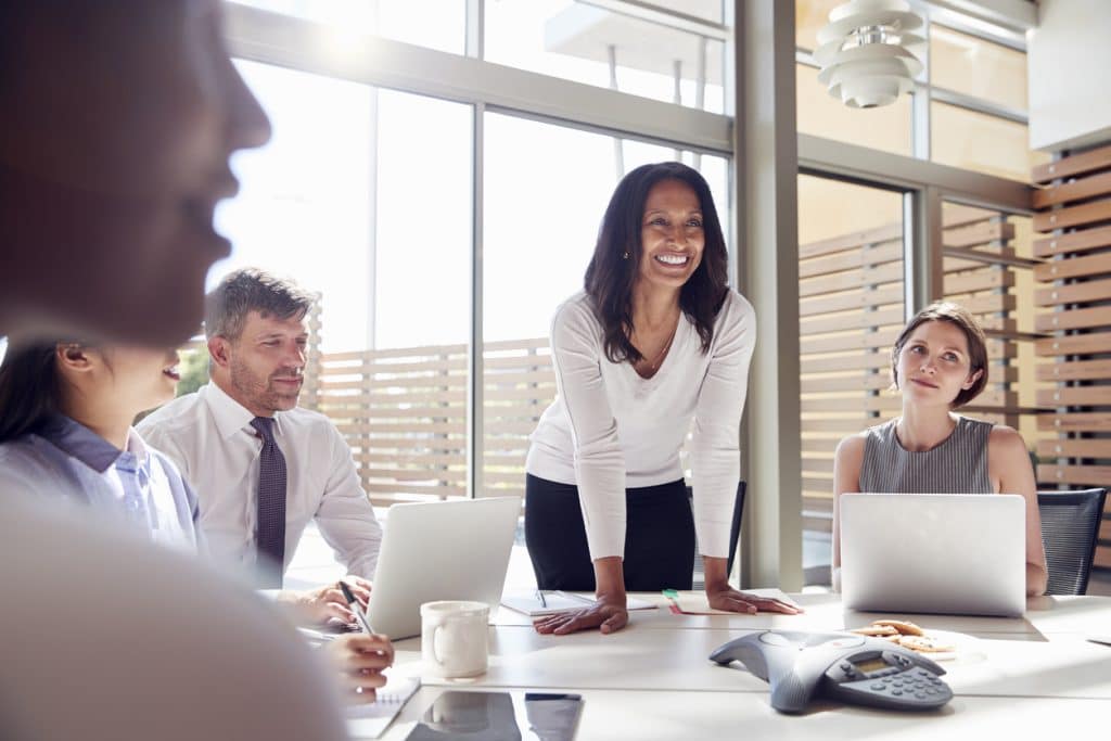 A woman is standing and smiling at the head of a meeting table, engaging with colleagues who are sitting with laptops open. Sunlight streams in through large windows in a modern office space, creating a bright and collaborative atmosphere—an ideal setting to discuss Customized Call Management Services.