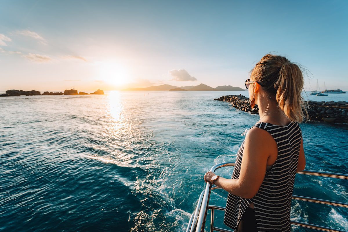 A woman with blonde hair in a ponytail stands on the deck of a boat, gazing at a beautiful sunset over the ocean. She wears a sleeveless striped top and sunglasses. The water sparkles in the sunlight, and distant mountains and other boats are visible on the horizon. Capturing this moment would be a breeze with iOS 18's new camera features.