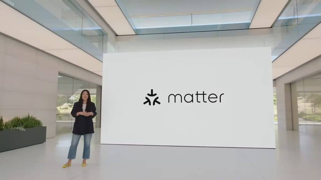 A woman stands in a modern, spacious presentation room next to a large screen displaying the logo and name "Matter." The room features sleek design elements with large windows and a glass structure overhead. Wearing a dark blazer, jeans, and yellow shoes, she presents on Matter's smart home device support.