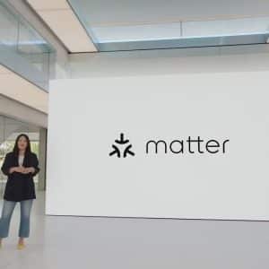 A woman stands in a modern, spacious presentation room next to a large screen displaying the logo and name "Matter." The room features sleek design elements with large windows and a glass structure overhead. Wearing a dark blazer, jeans, and yellow shoes, she presents on Matter's smart home device support.