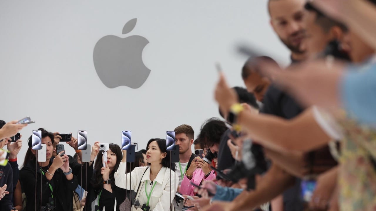 A group of people, many holding smartphones, gather to photograph and record new iPhones displayed vertically. The large Apple logo is visible in the background, suggesting this is an Apple event. With the crowd excited and engaged, it's clear why Apple's Q2 2024 earnings surpasses analyst expectations.