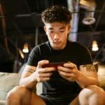 A young man sits on a couch, intently playing Valorant Mobile on his smartphone. He is wearing a black T-shirt and shorts, with tattoos visible on his arms. The background is dimly lit with industrial-style ceiling lights, adding to the intense atmosphere as he masters tips for beginners.