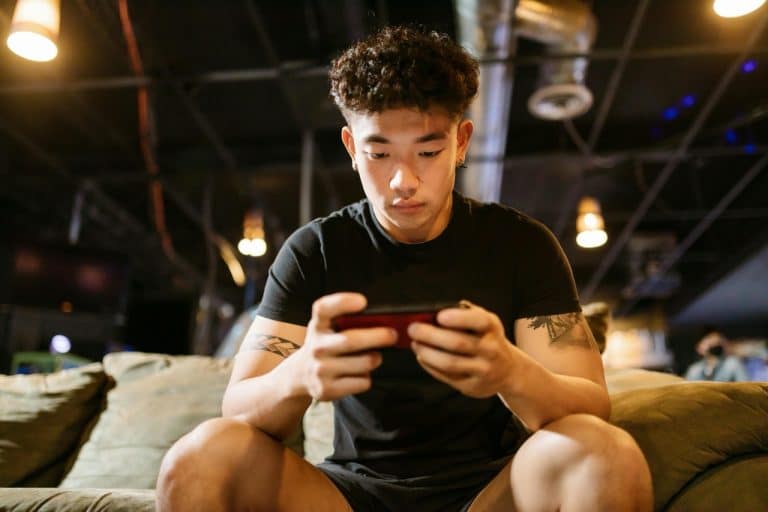 A young man sits on a couch, intently playing Valorant Mobile on his smartphone. He is wearing a black T-shirt and shorts, with tattoos visible on his arms. The background is dimly lit with industrial-style ceiling lights, adding to the intense atmosphere as he masters tips for beginners.