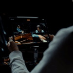 A person holds an iPad displaying a video with four split-screen views: an overhead shot, a side view, a close-up of hands on a piano, and a person playing the piano. The background is dark, highlighting the bright screen of the tablet showcasing the impressive Multicam Feature in Final Cut Pro.