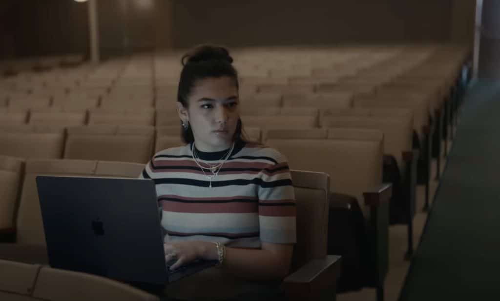 A woman with a striped shirt sits alone in an empty auditorium, working on a laptop. She has dark hair tied up and wears multiple necklaces and bracelets. The dim lighting highlights her focused expression as she benefits from impressive Mac battery life.