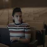 A woman with a striped shirt sits alone in an empty auditorium, working on a laptop. She has dark hair tied up and wears multiple necklaces and bracelets. The dim lighting highlights her focused expression as she benefits from impressive Mac battery life.