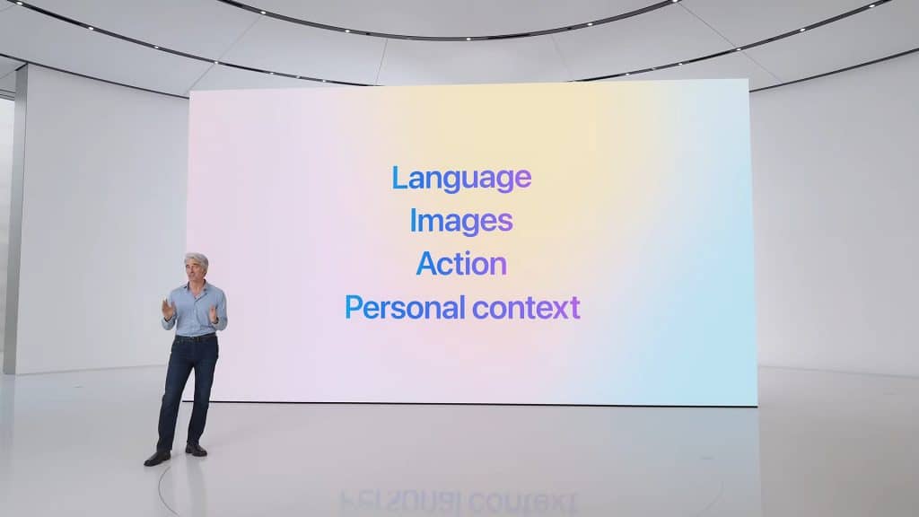 A person stands on a minimalist stage with a large screen behind them. The screen, displaying the text "Language," "Images," "Action," and "Personal context" in blue on a gradient background, hints at themes related to Apple Intelligence Plans. The individual appears to be giving a presentation.