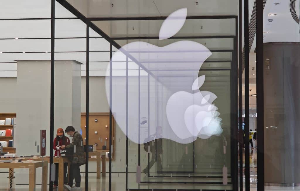 A large, illuminated Apple logo is displayed on the glass facade of an Apple Store. Inside the store, two people are seen browsing and interacting with various Apple products on display tables. The store features a minimalist design with wooden tables and a bright interior showcasing the latest in watchOS 11 technology.
