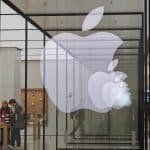 A large, illuminated Apple logo is displayed on the glass facade of an Apple Store. Inside the store, two people are seen browsing and interacting with various Apple products on display tables. The store features a minimalist design with wooden tables and a bright interior showcasing the latest in watchOS 11 technology.