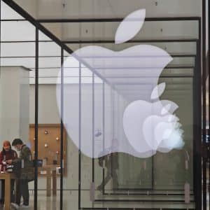 A large, illuminated Apple logo is displayed on the glass facade of an Apple Store. Inside the store, two people are seen browsing and interacting with various Apple products on display tables. The store features a minimalist design with wooden tables and a bright interior showcasing the latest in watchOS 11 technology.