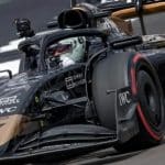 A Formula 1 race car drives on a track during a race. The vehicle features black and gold accents and sponsorship logos such as IWC and Express. The driver, wearing a helmet and visor, is focused ahead, while there's a camera mounted on top of the car, reminiscent of scenes from an Apple F1 movie set for a 2025 release.