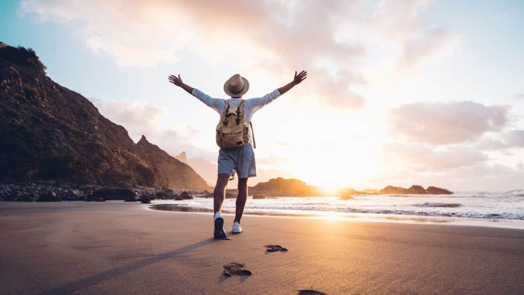 A person wearing a hat and a backpack walks along a beach at sunset with arms outstretched. Footprints trail behind them on the sandy shore, and rocky cliffs rise on the left. The ocean waves gently lap at the shore under a partly cloudy sky—an ideal moment to capture using one of the top travel apps.