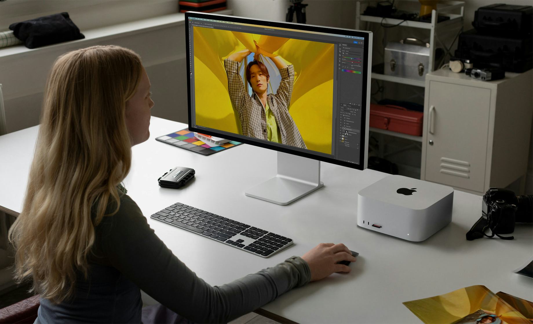 M4 Mac Upgrades: A person with long hair is sitting at a desk, working on Adobe photo editing software on a large computer monitor. The displayed image shows a person posing with their arms raised against a yellow backdrop. The desk is equipped with a keyboard, mouse, and various devices, adhering to Adobe subscription practices.