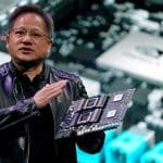 A person wearing a black leather jacket holds up a circuit board with two large chips, speaking in front of a projected background image that features a close-up of computer hardware components, resembling the innovation seen in companies like Nvidia.