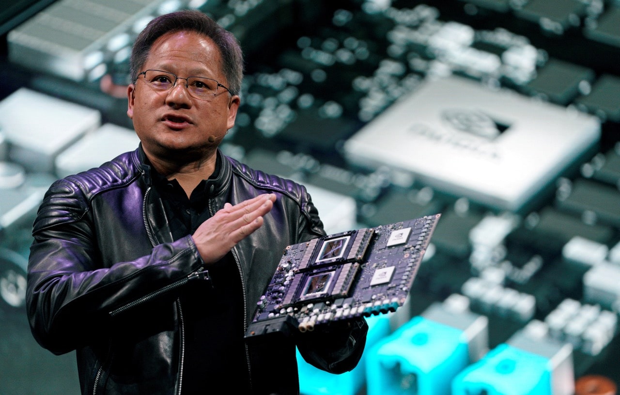 A person wearing a black leather jacket holds up a circuit board with two large chips, speaking in front of a projected background image that features a close-up of computer hardware components, resembling the innovation seen in companies like Nvidia.