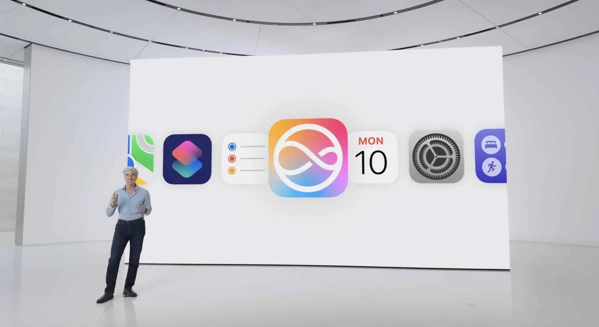 Person presenting on stage with a large screen displaying various app icons, including a calendar set to Monday the 10th in the center. The setting appears to be a modern, white, minimalist design with curved architectural elements and sleek Apple devices scattered around.