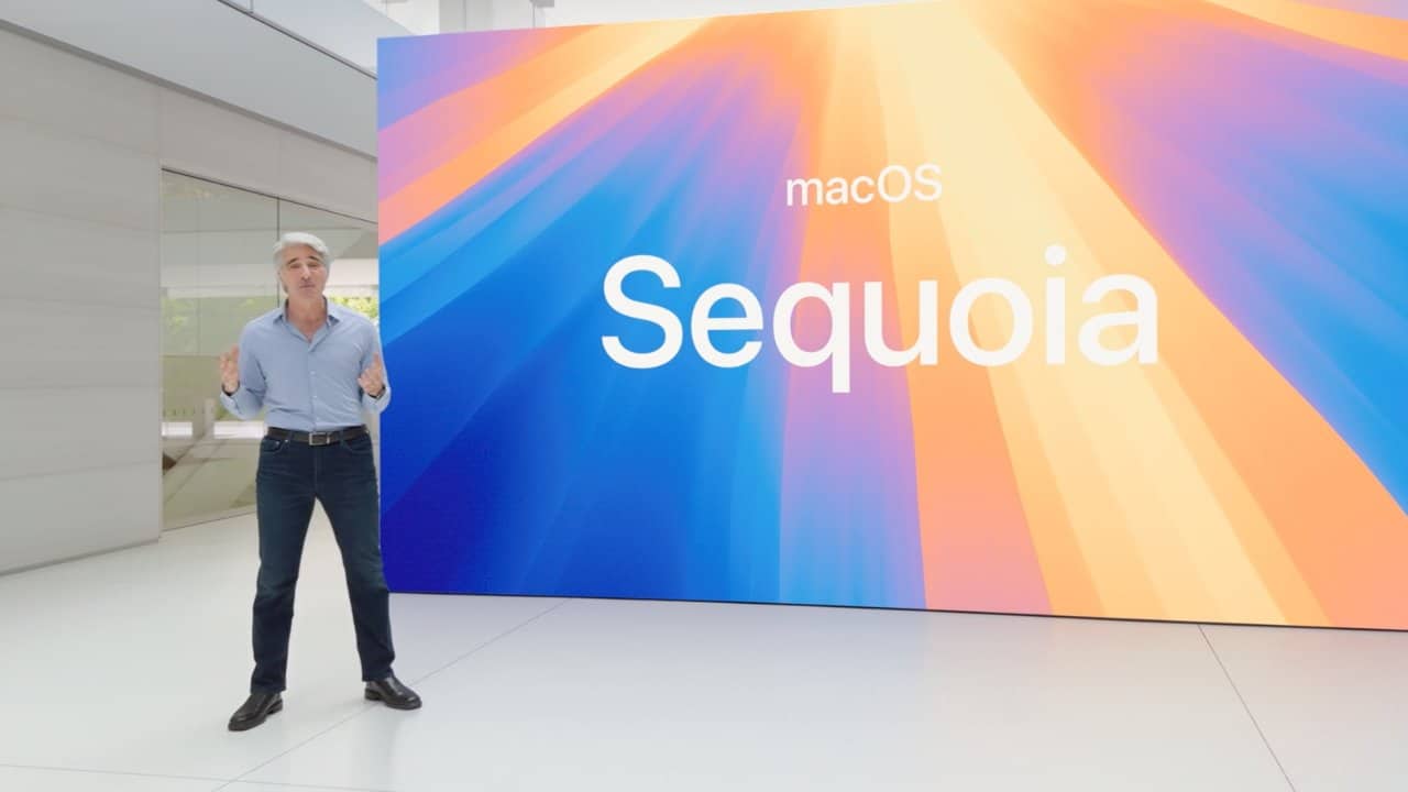 A person stands near a large screen displaying "macOS Sequoia" with vibrant colors of blue, orange, and yellow rays. The individual appears to be giving a presentation or announcement. The surroundings are modern with a minimalist design.