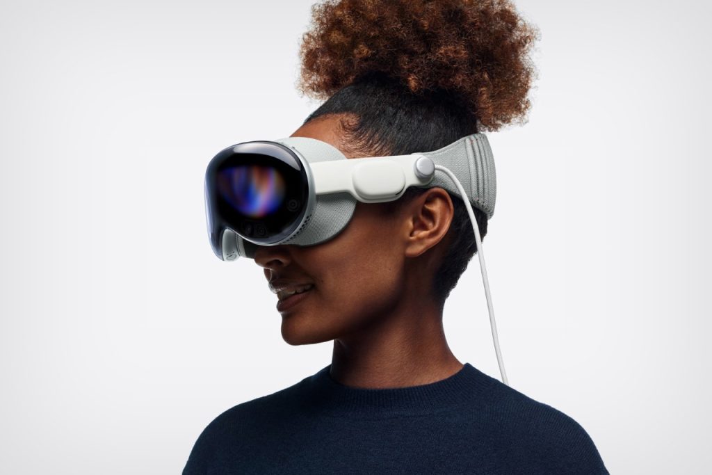 A person with curly hair in a bun wears a sleek, futuristic virtual reality headset with a white strap. The person is smiling and looking to the side, embodying tech innovations with a plain white background behind them.