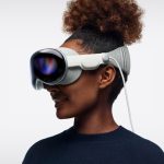 A person with curly hair in a bun wears a sleek, futuristic virtual reality headset with a white strap. The person is smiling and looking to the side, embodying tech innovations with a plain white background behind them.