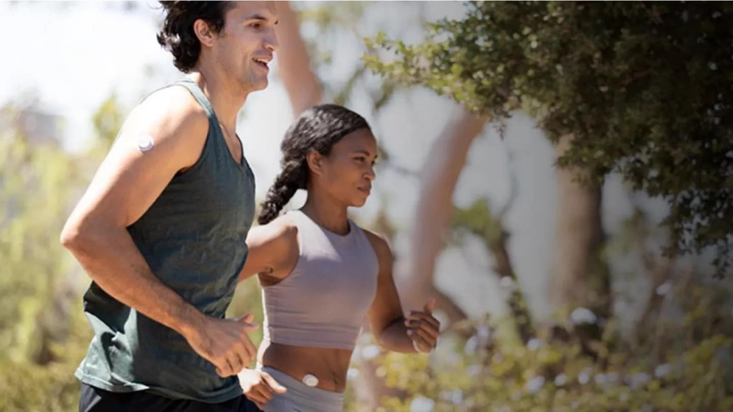 A man and a woman are jogging outdoors on a sunny day, surrounded by trees. Both are smiling and wearing fitness attire, including sleeveless tops. They each have a small round Dexcom Sensor on their upper arms, suggesting medical or health monitoring devices.