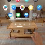 A person is using an augmented reality headset in a living room with wooden furniture and a patterned rug. The Apple Intelligence AR interface displays various app icons like Mail, Photos, and App Store floating in the air before them.