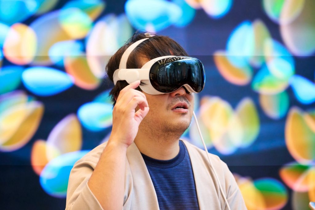 A person with dark hair is wearing an Apple In-Store Vision Pro headset and gesturing with one hand. The background features colorful, out-of-focus lights. The individual appears to be deeply engaged with the VR content and exploring its new features.