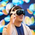 A person with dark hair is wearing an Apple In-Store Vision Pro headset and gesturing with one hand. The background features colorful, out-of-focus lights. The individual appears to be deeply engaged with the VR content and exploring its new features. | Apple Vision Pro international expansion delivers augmented reality technology to new global markets