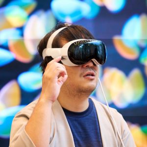 A person with dark hair is wearing an Apple In-Store Vision Pro headset and gesturing with one hand. The background features colorful, out-of-focus lights. The individual appears to be deeply engaged with the VR content and exploring its new features.