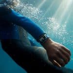 Person wearing a wetsuit and an Apple Watch, swimming underwater with sunlight filtering through the water surface. Bubbles surround the swimmer, and the smartwatch display is visible on their wrist as they use Find My to locate a missing item.