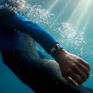 Person wearing a wetsuit and an Apple Watch, swimming underwater with sunlight filtering through the water surface. Bubbles surround the swimmer, and the smartwatch display is visible on their wrist as they use Find My to locate a missing item.