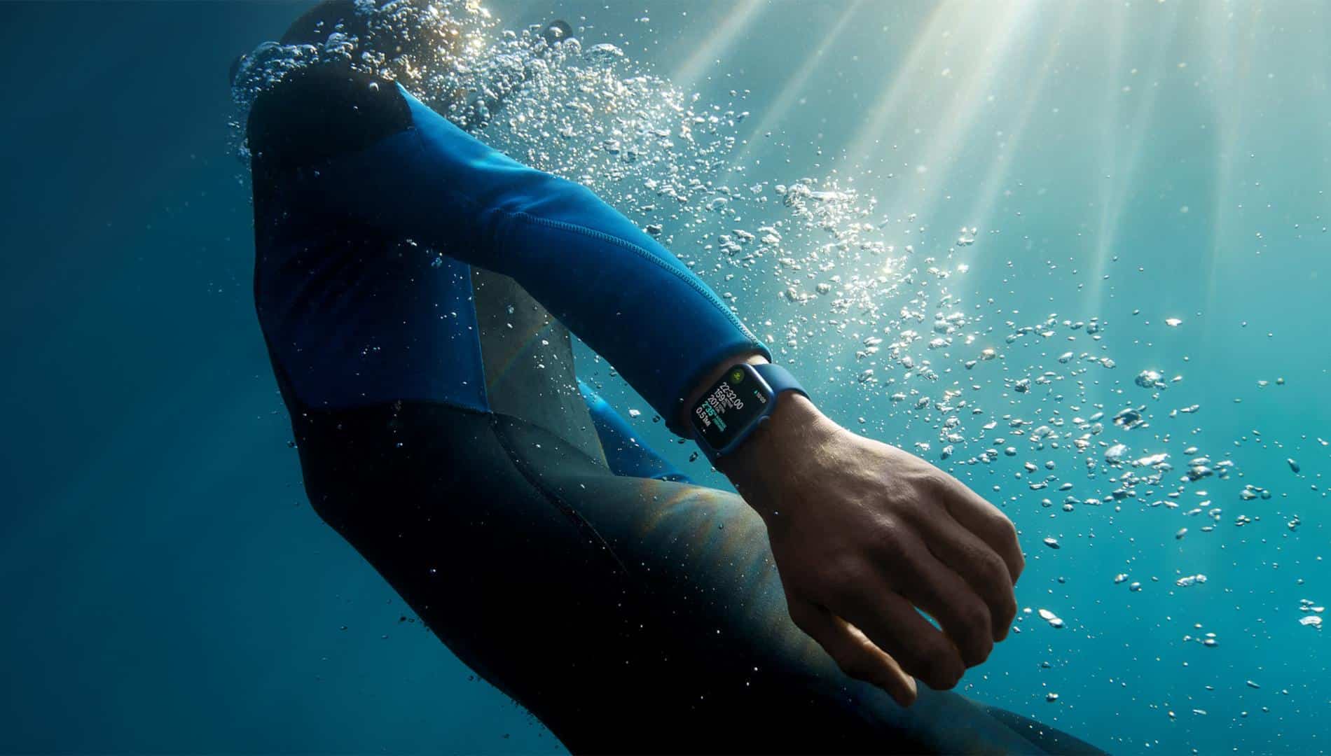 Person wearing a wetsuit and an Apple Watch, swimming underwater with sunlight filtering through the water surface. Bubbles surround the swimmer, and the smartwatch display is visible on their wrist as they use Find My to locate a missing item.