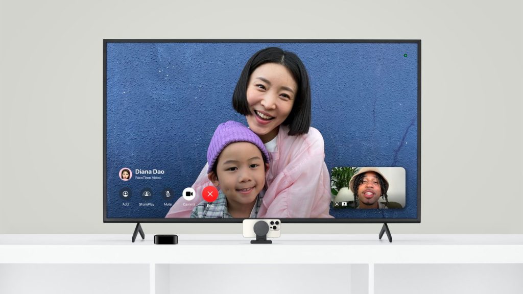 tvOS 18: A video call is displayed on a large TV screen via Apple TV. A woman and a child are smiling and looking at the camera, with a blue wall in the background. Inset at the bottom right, another person appears on FaceTime. The devices for the call are on a white table below the TV.