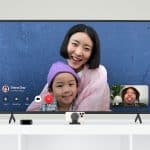 tvOS 18: A video call is displayed on a large TV screen via Apple TV. A woman and a child are smiling and looking at the camera, with a blue wall in the background. Inset at the bottom right, another person appears on FaceTime. The devices for the call are on a white table below the TV.