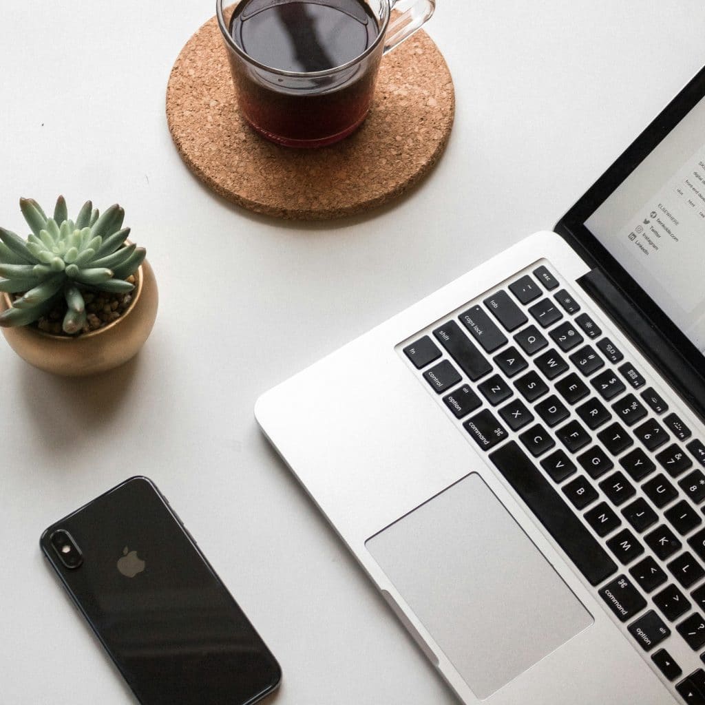 A laptop with an open screen sits on a white desk next to a smartphone, a cup of black coffee on a cork coaster, and a small potted succulent plant. The minimalist workspace is ideal for tackling tasks like adjusting network settings in Apple Mail.