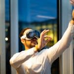 A person wearing a virtual reality headset gestures with one hand raised and another forming a pinch motion. They are indoors, standing near large, vertical windows, and are dressed in a white long-sleeve shirt. Engrossed in their VR experience, they may be curious about recent IDC Reports on US Vision Pro Sales decline.