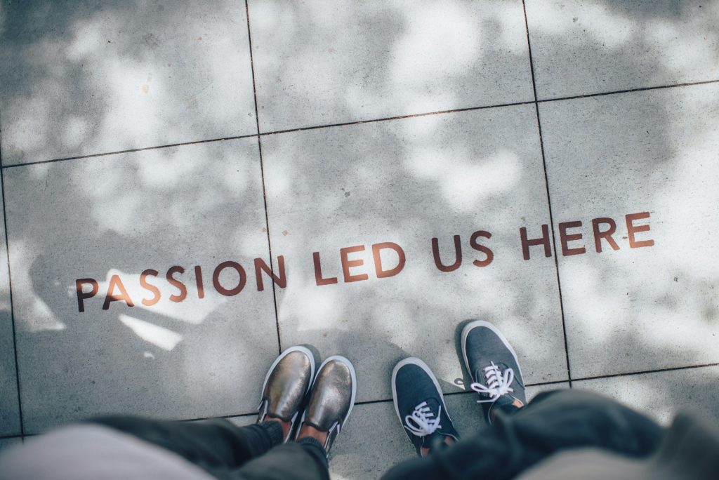 Two people standing on a paved surface with the words "PASSION LED US HERE" inscribed on the ground between them. The individuals are wearing sneakers, one pair silver and the other pair blue. Dappled sunlight and tree shadows are visible on the pavement, creating a whimsical snowflake pattern.