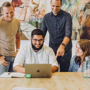 Four people are gathered around a table with laptops, smiling and engaging in conversation about ETL processes. The group consists of three men and one woman. One man is pointing at a laptop screen displaying Google Ads data while others look on. The background features colorful abstract artwork.