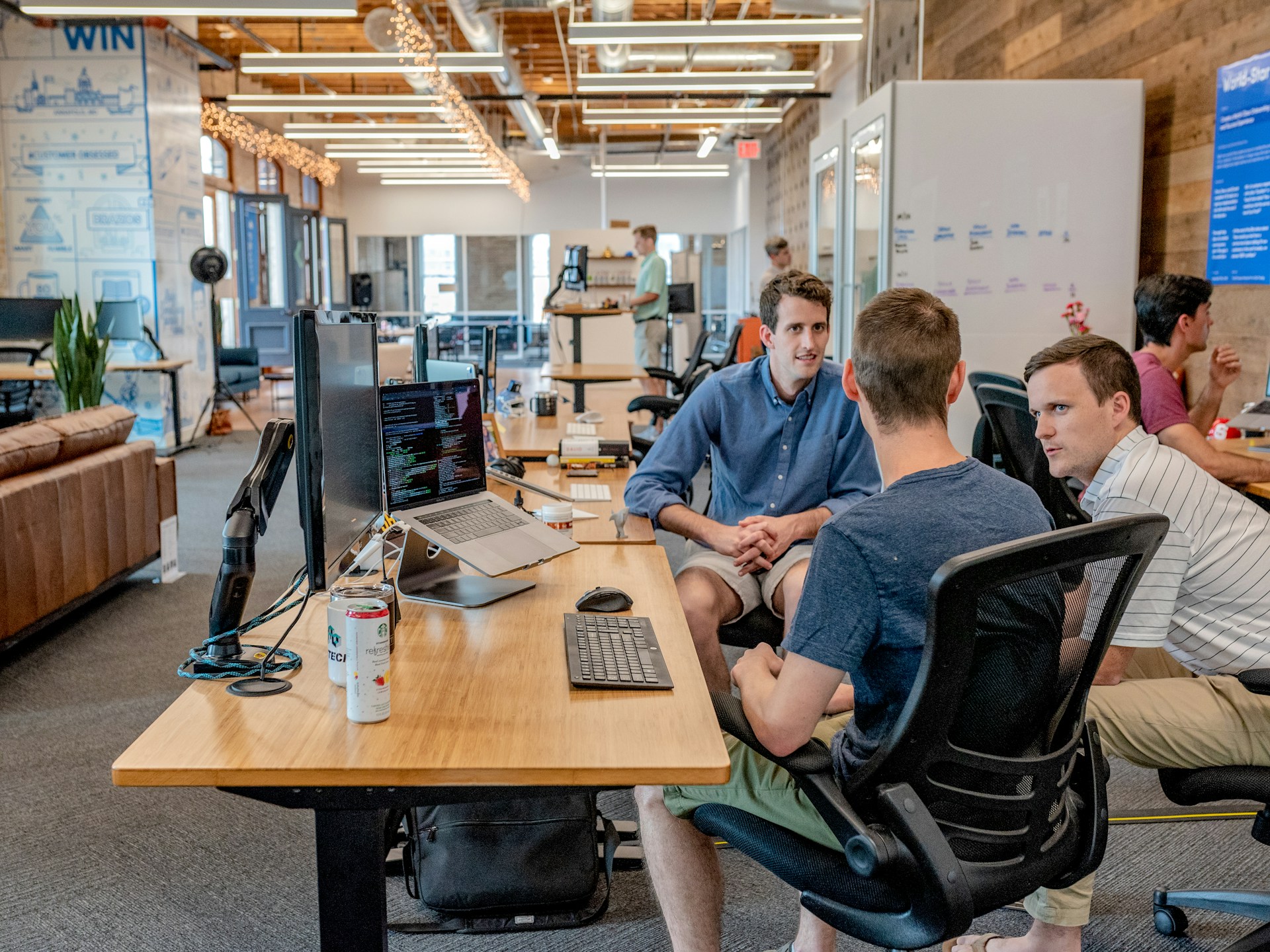 A group of three people sitting around a desk in a modern, open-plan office, collaborating and discussing Google Ads on a computer screen. The office space features wooden walls, multiple computer stations, exposed ceilings, and casual seating areas.