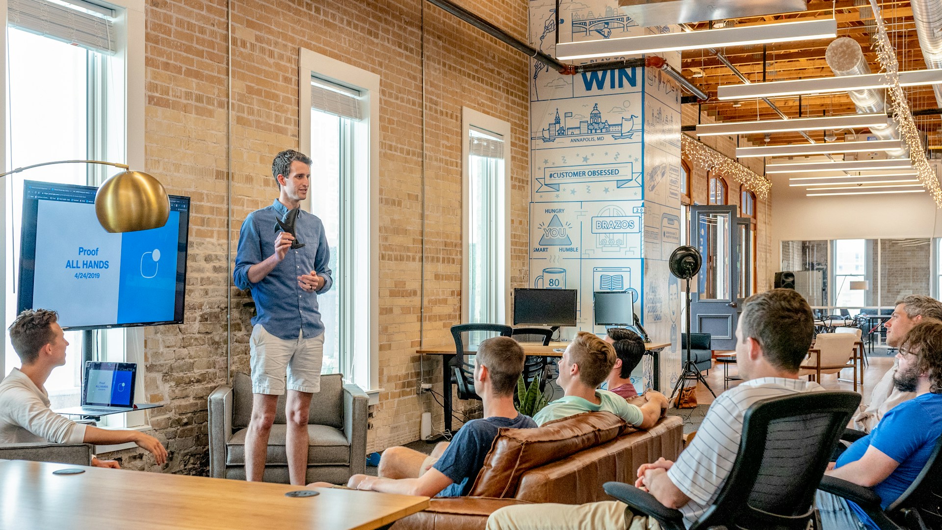 A man stands at the front of a modern office space giving a presentation to colleagues about an advanced ETL process. He is speaking and gesturing towards a screen displaying a slide titled "Proof ALL HANDS 04/2019." The room has brick walls, large windows, and a casual setup.