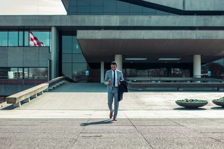 A man in a gray suit walks confidently while holding a phone and jacket in front of a modern building with large glass windows and concrete elements. There is a distant national flag on the left, two circular flower planters on the right, and an ad for Google Ads displayed prominently.