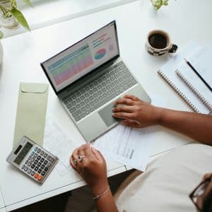 A person is working on a laptop at a desk, analyzing charts and graphs related to Google Ads. The desk also has a calculator, a cup of coffee, an envelope, and a notebook with a pencil. Several plants are placed around the workspace, creating a serene environment for their ETL tasks.