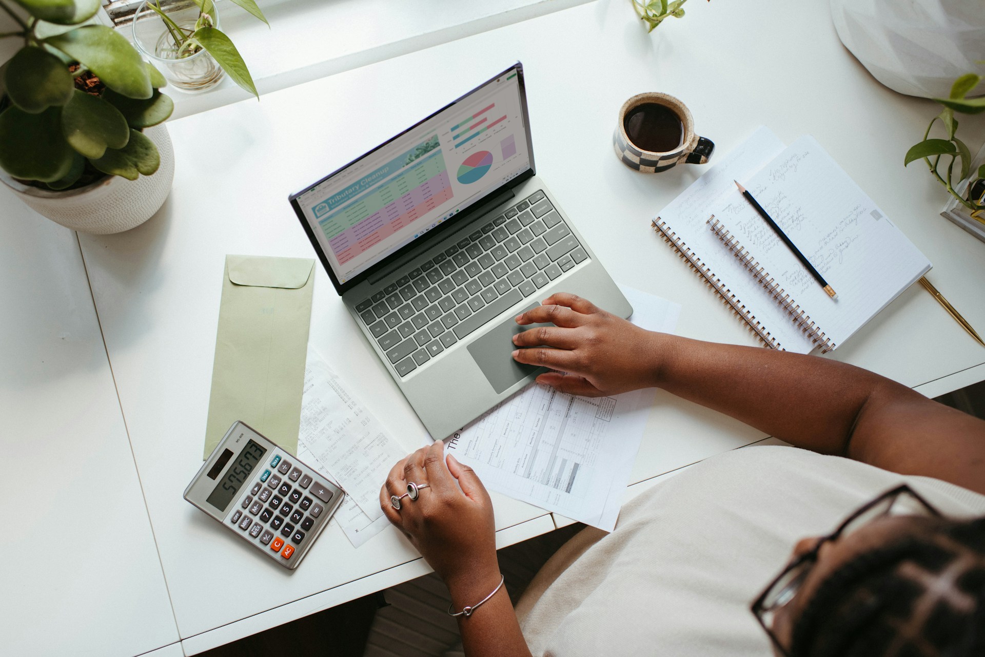 A person is working on a laptop at a desk, analyzing charts and graphs related to Google Ads. The desk also has a calculator, a cup of coffee, an envelope, and a notebook with a pencil. Several plants are placed around the workspace, creating a serene environment for their ETL tasks.