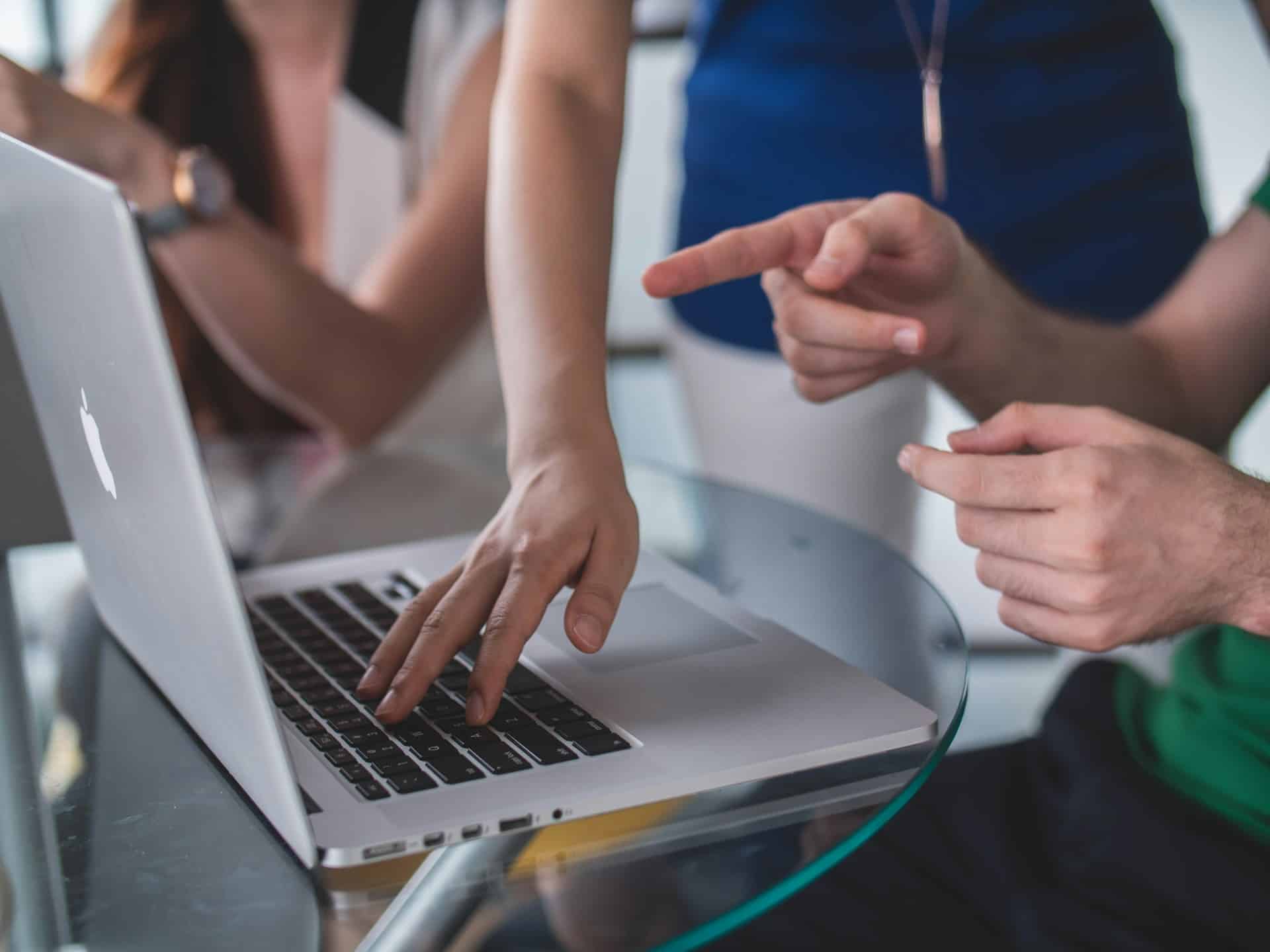 Three individuals are gathered around an open laptop on a glass table. One person is typing on the keyboard, while another points towards the screen, possibly discussing their Google Ads campaign. The third person is partially visible, also focused on the laptop. The background is slightly blurred.