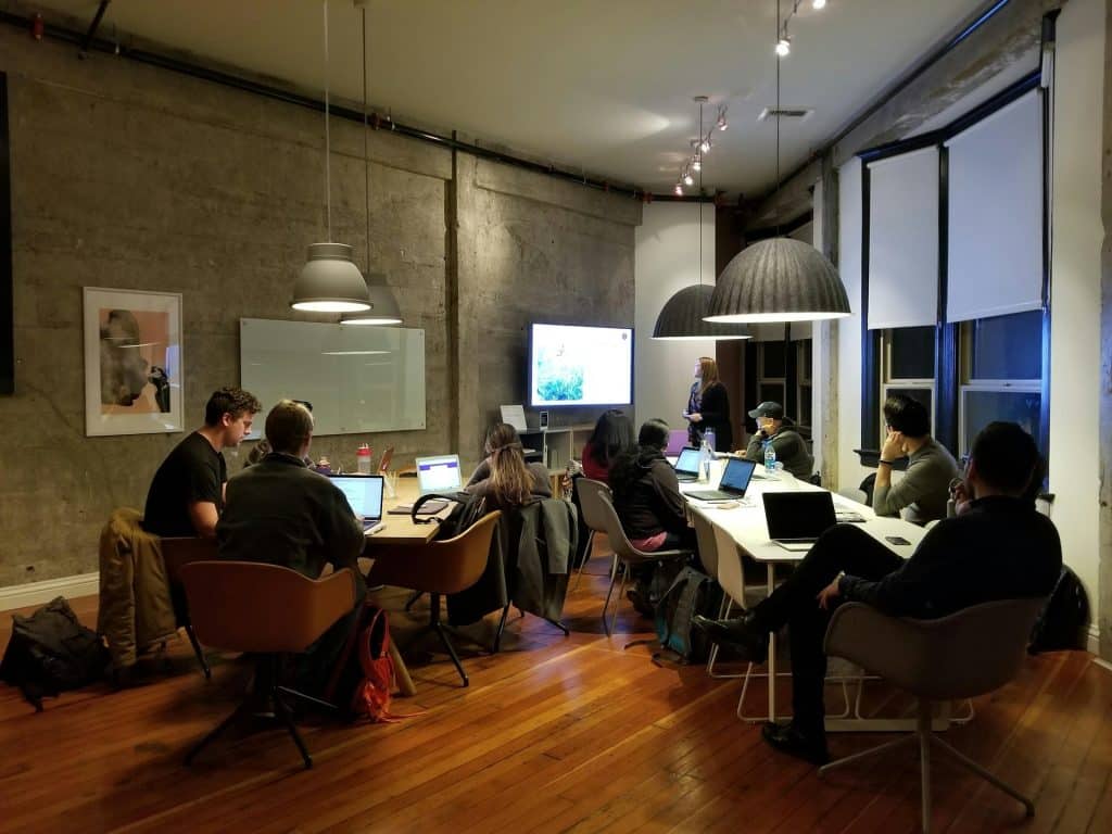 A group of people is sitting around tables in a modern office setting, working on laptops and other devices. A person stands at the front near a large screen displaying information about ETL in Marketing. The room has a polished wood floor, pendant lights, and large windows with shades.